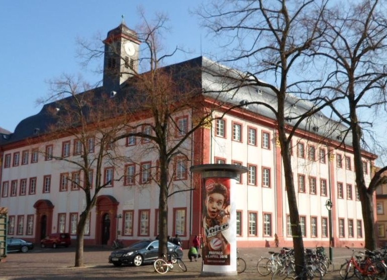 Historisches rot-weißes Gebäude mit Uhrturm in der Heidelberger Altstadt, davor Bäume, Fahrräder und eine Litfaßsäule