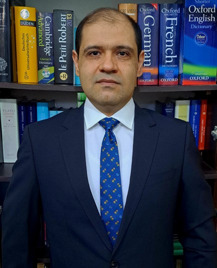 Dr. Mahdi Kohandani im dunklen Anzug mit blauer Krawatte vor einem Bücherregal mit Wörterbüchern